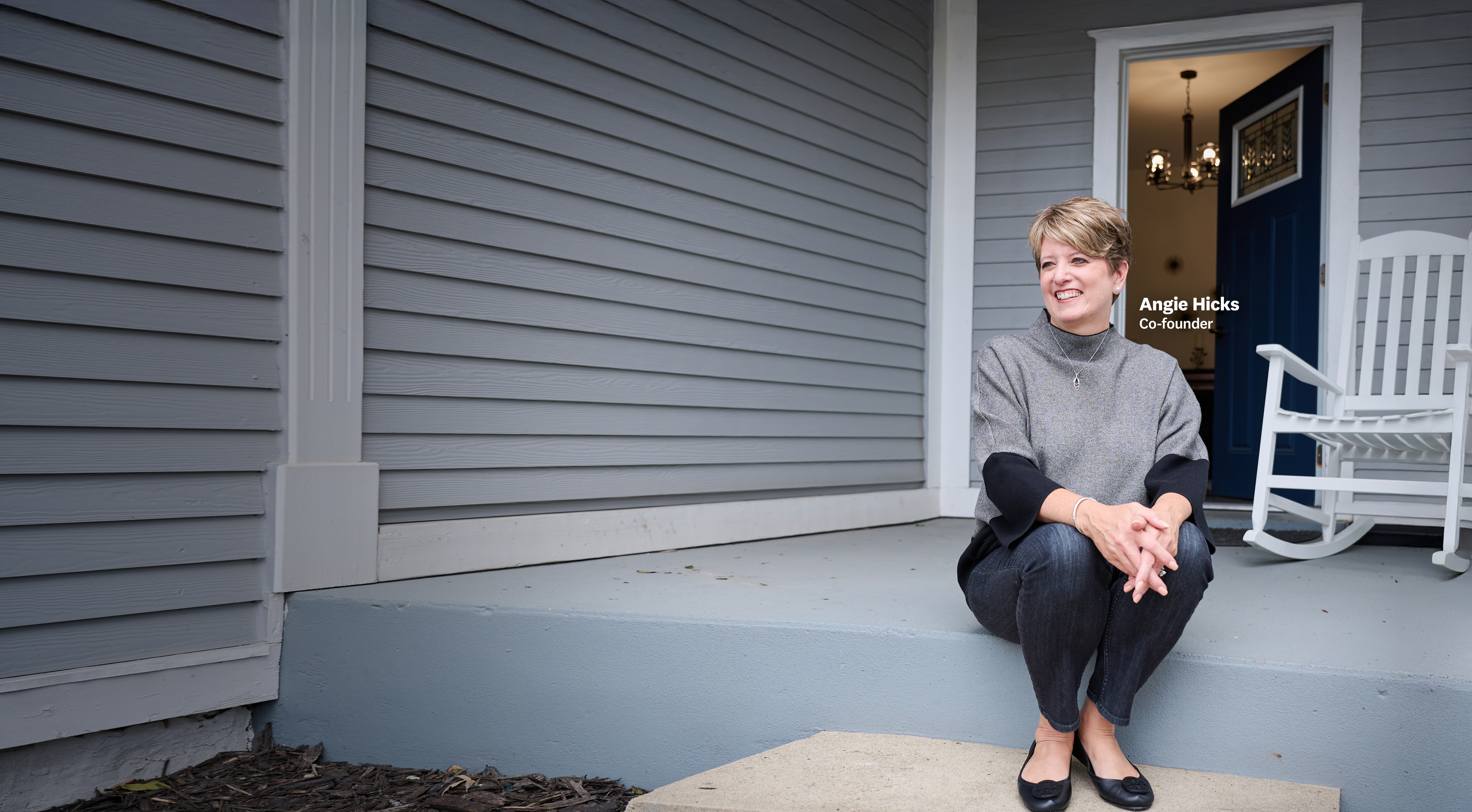 Angie Hicks sitting on the front porch steps of a gray house, smiling, with a blue door and a white rocking chair visible in the background.